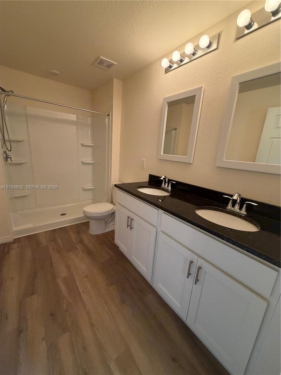 Dual-sink vanity with dark stone-finish countertop, white cabinetry, wood-finish flooring, and a shower-tub combination featuring built-in shelving