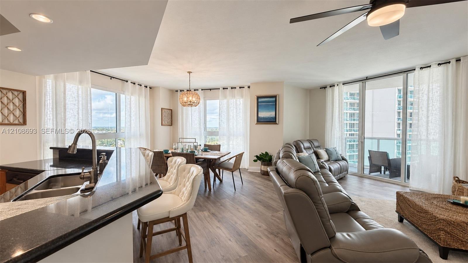Open-concept interior featuring wood-finish flooring, a kitchen peninsula with a stone countertop, an integrated stainless steel dual-basin sink, floor-to-ceiling glass panel doors, and a modern drum-style pendant light