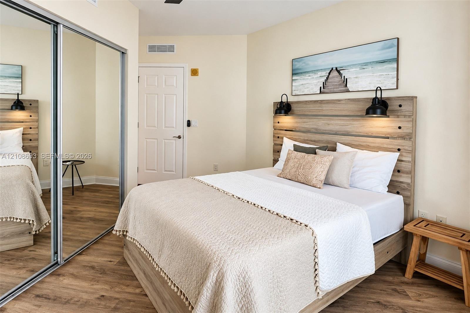 Wood-finish flooring, full-height mirrored sliding closet doors, integrated wood-tone headboard, and dual wall-mounted light fixtures