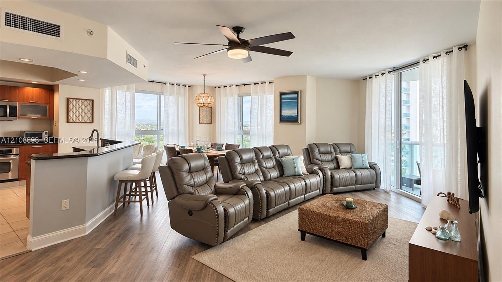 Open-concept area featuring wood-finish flooring, a kitchen breakfast bar with dark countertops, floor-to-ceiling windows, and a central ceiling fan