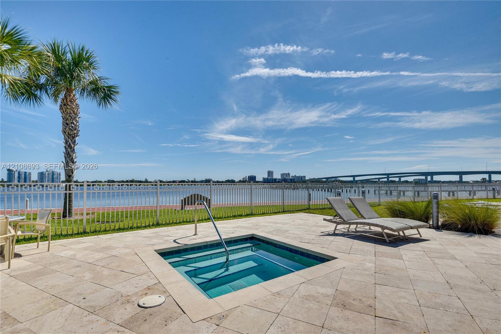 In-ground spa with a metal handrail, stone tile deck, and metal perimeter fencing overlooking a waterfront bulkhead