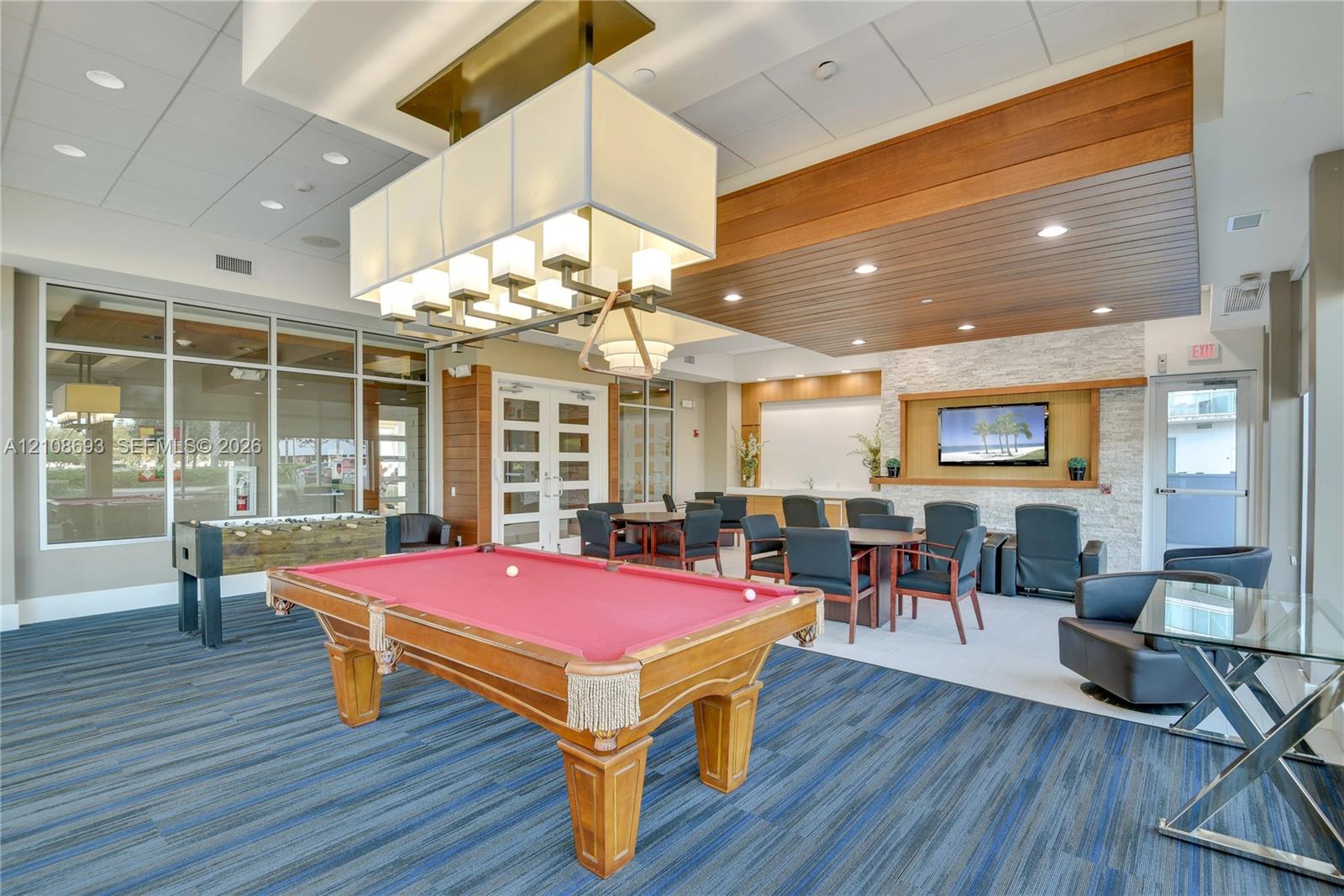 Recreation space featuring a coffered ceiling with integrated wood paneling, stone accent wall, large-scale box chandelier, and expansive floor-to-ceiling windows