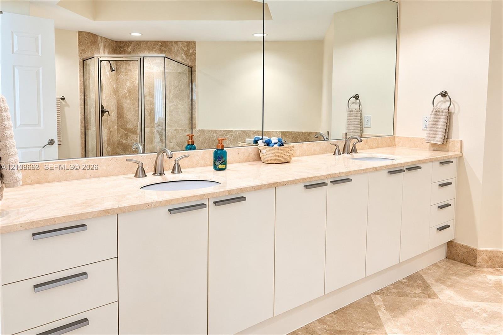 Extended dual-sink vanity featuring white flat-panel cabinetry, stone countertops, integrated sink basins, and polished metal hardware, accompanied by a large wall-mounted mirror, recessed lighting, and a walk-in shower with natural stone tile surrounds