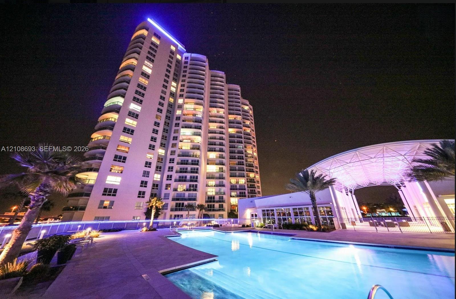 High-rise residential tower featuring tiered balconies, a sprawling swimming pool, expansive pool deck, and a covered pavilion with structural framework