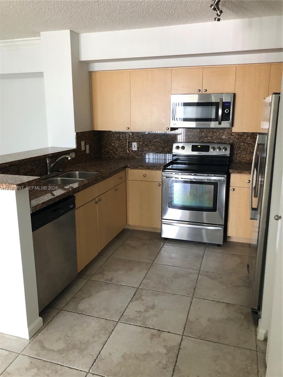 Kitchen featuring light wood-finish cabinetry, stone-finish countertops, stainless steel appliances, a double-basin sink, and tile flooring