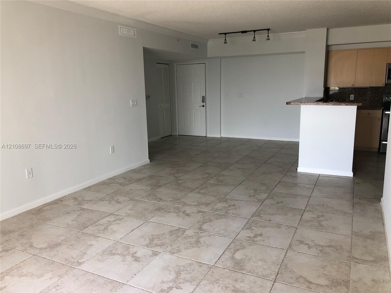 Open living area featuring neutral tile flooring, track lighting, and a kitchen peninsula with a stone-finish countertop and wood cabinetry