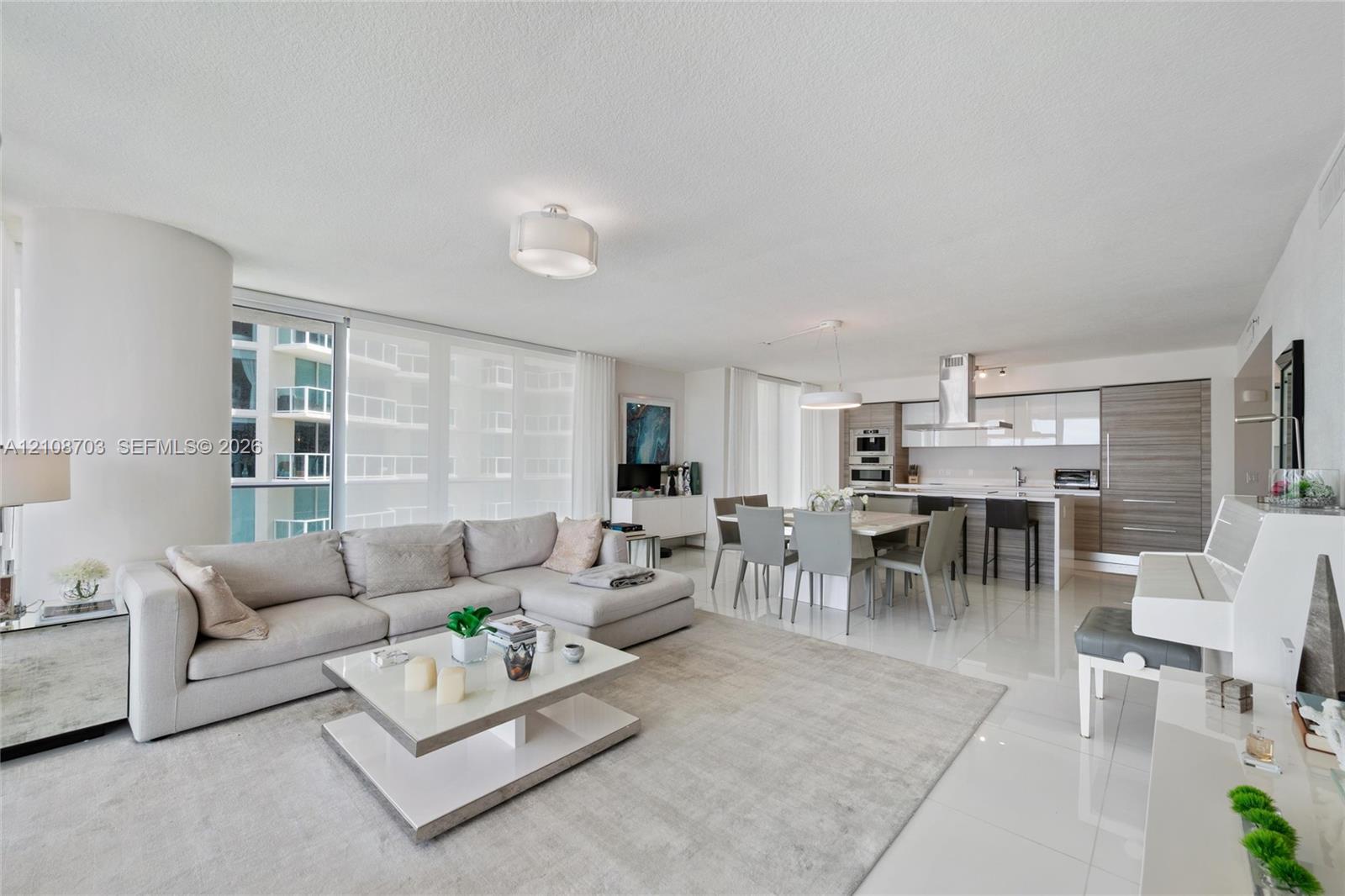 Open-concept living area featuring large-format tile flooring, floor-to-ceiling windows, modern cabinetry with wood-finish and high-gloss accents, and stainless steel built-in appliances