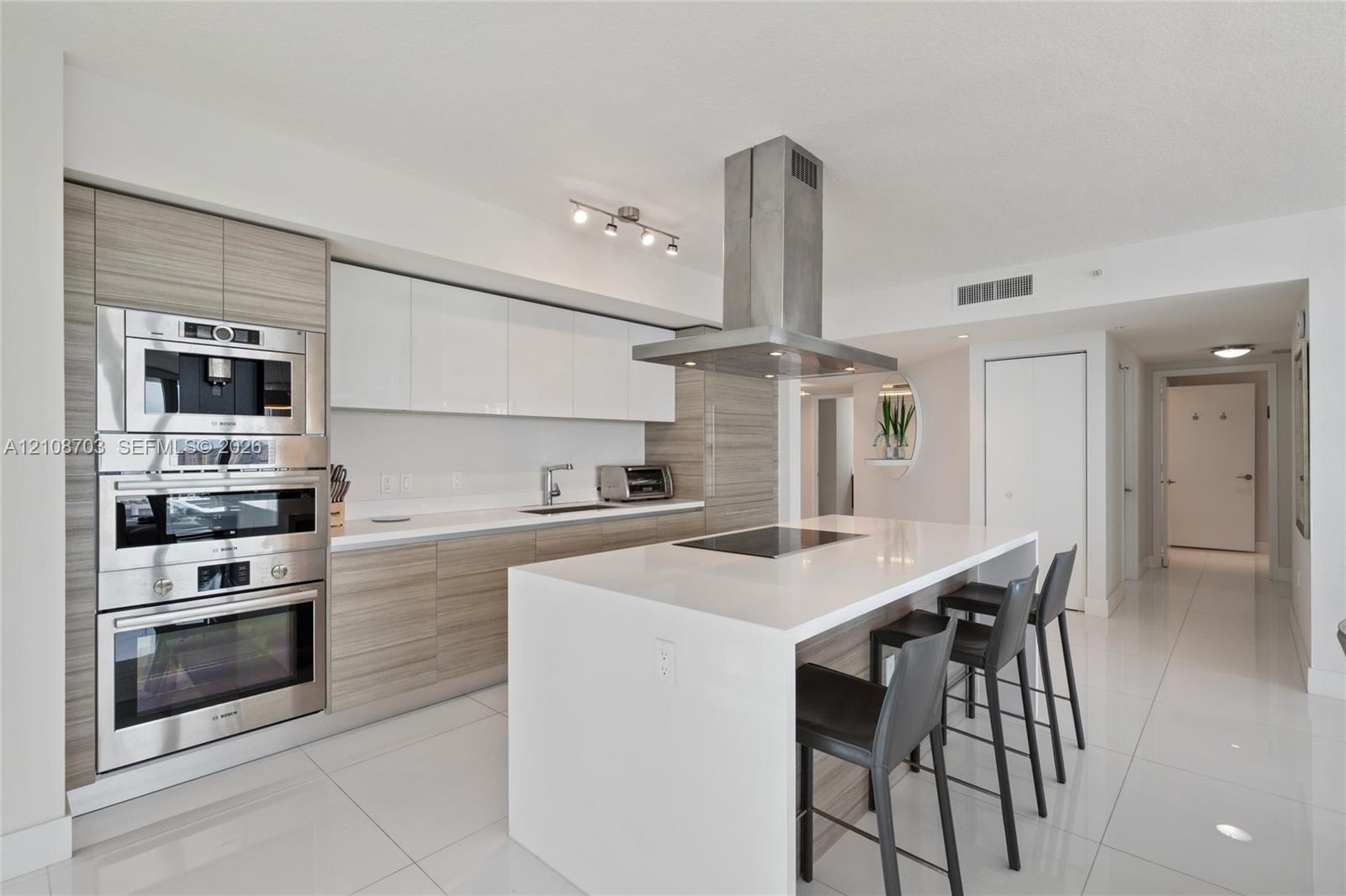 Kitchen featuring a central island with a solid-surface countertop, high-gloss white and wood-finish cabinetry, built-in stainless steel appliances, and a ceiling-mounted stainless steel range hood