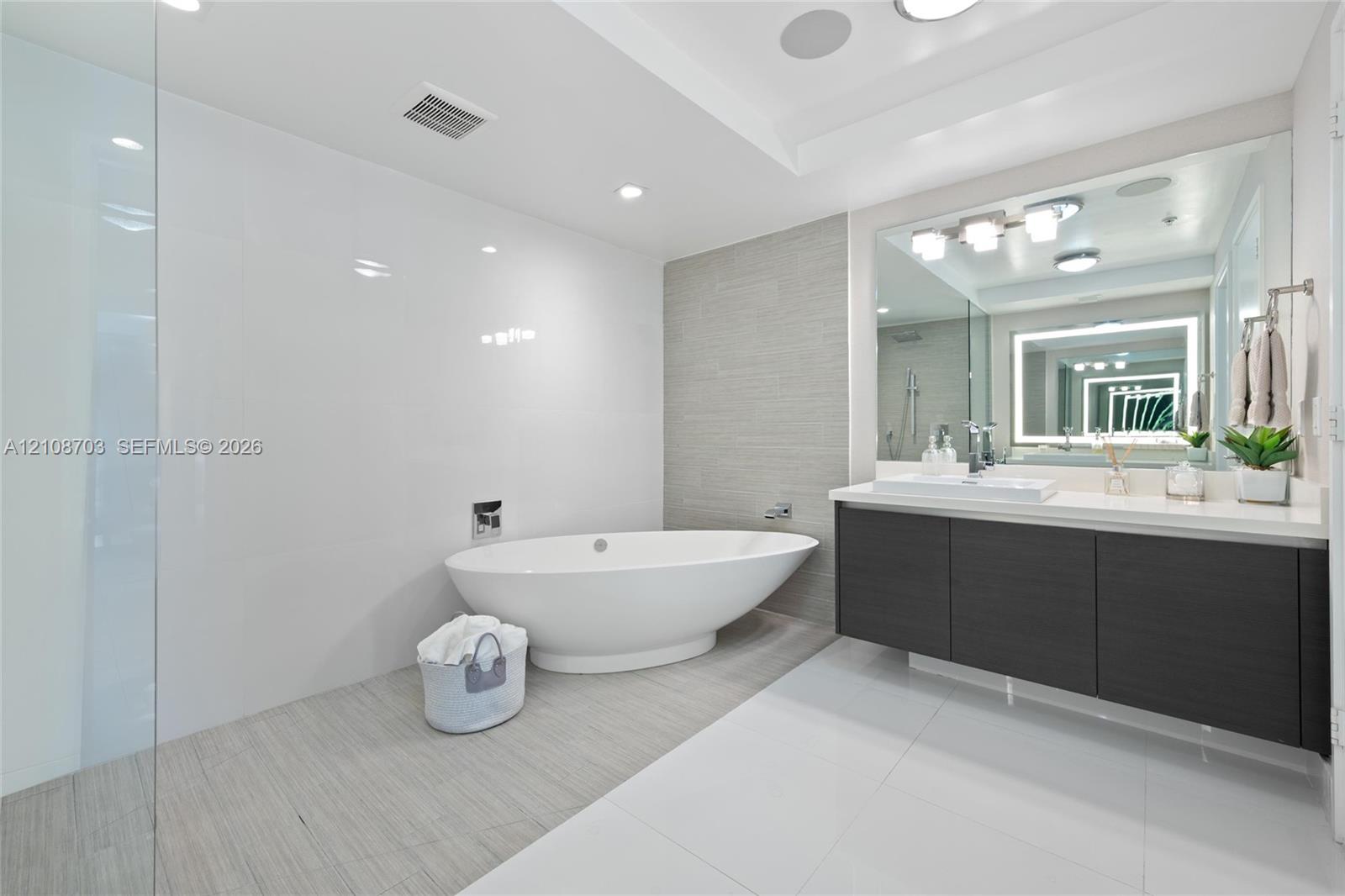 Freestanding soaking tub, walk-in glass shower, wall-mounted vanity with integrated sink, large-format polished tile flooring, and textured tile accent wall