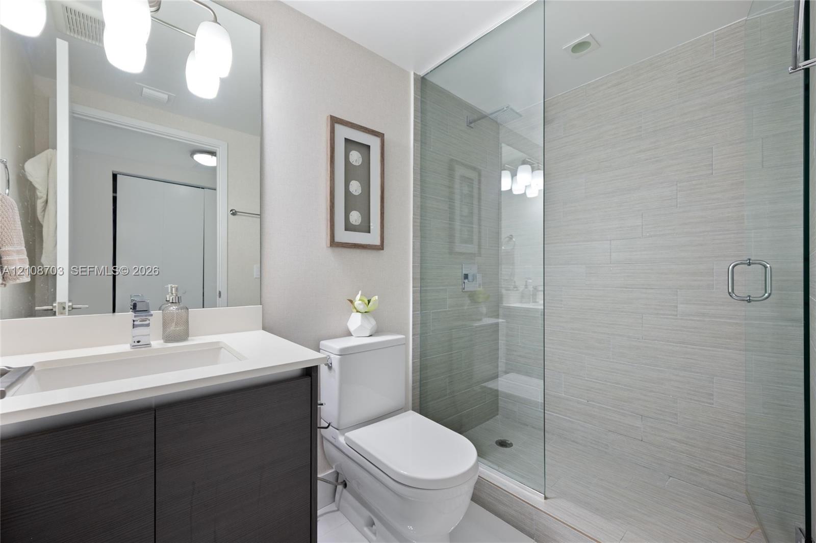Bathroom featuring a dark wood-finish vanity with a white countertop, integrated sink, large wall-mounted mirror, and walk-in shower with floor-to-ceiling tile and a frameless glass door