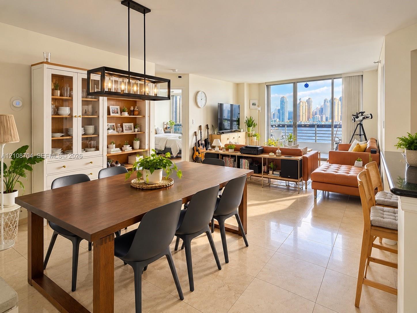 Open-concept floor plan featuring large-format tile flooring, built-in floor-to-ceiling cabinetry with glass-paneled display doors, a linear industrial-style pendant light fixture, and floor-to-ceiling windows with waterfront and city skyline scenery Open-concept floor plan featuring large-format tile flooring, built-in floor-to-ceiling cabinetry with glass-paneled display doors, a linear industrial-style pendant light fixture, and floor-to-ceiling windows with waterfront and city skyline scenery