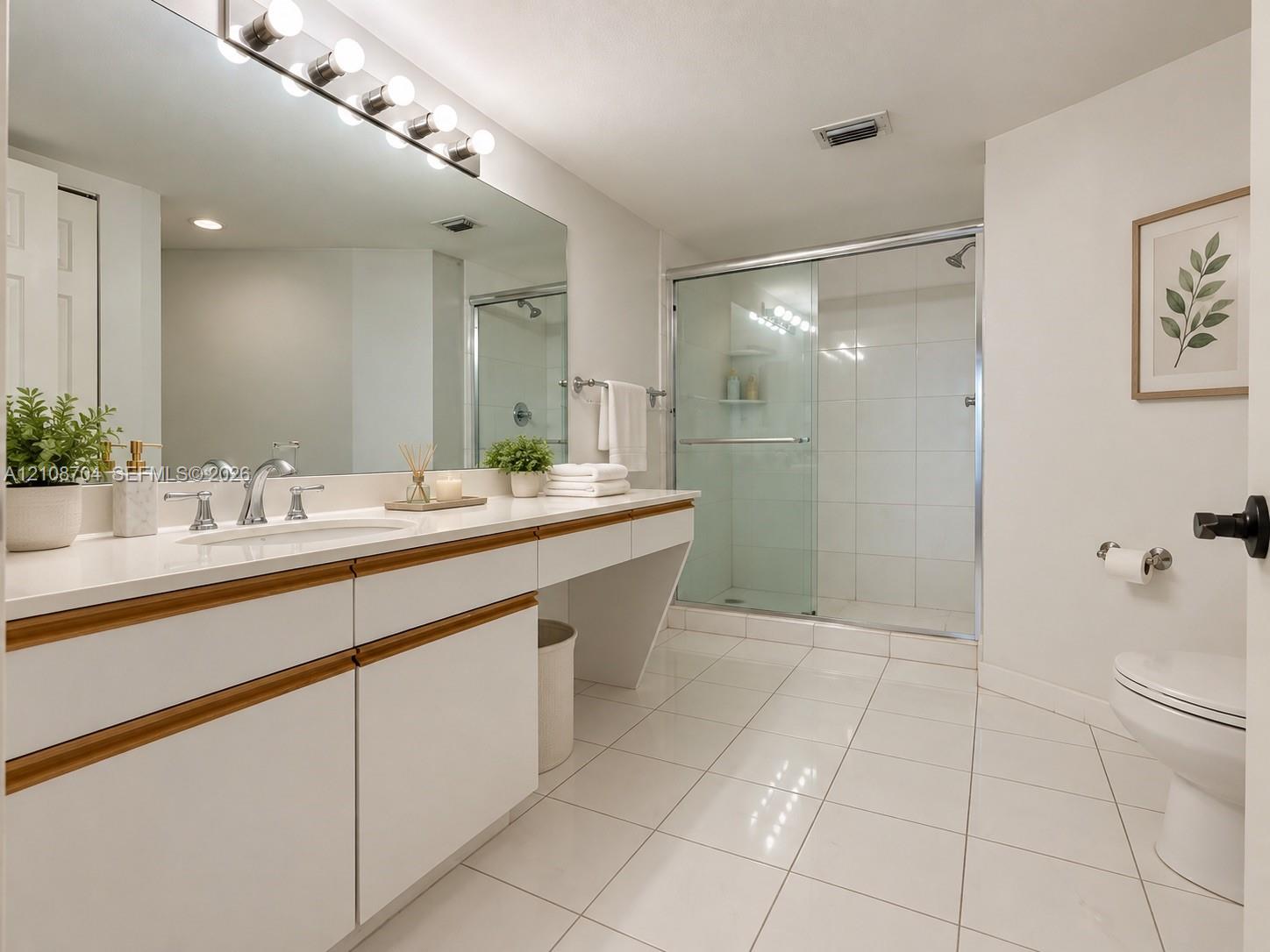 Bathroom featuring a dual-tone vanity with a solid-surface countertop, an expansive wall-to-wall mirror, porcelain tile flooring, and a walk-in shower enclosed by sliding glass doors