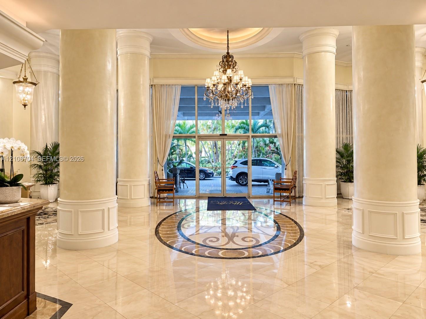 Grand lobby entrance featuring polished stone flooring with an inlaid mosaic medallion, structural fluted columns with detailed molding, a recessed circular ceiling medallion, and an ornate crystal chandelier