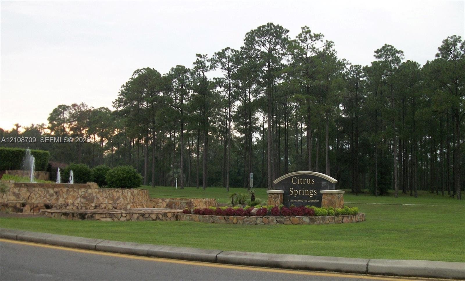 Community entrance featuring stone-veneer retaining walls, tiered landscaping, a multi-tiered fountain, and established pine forest surroundings