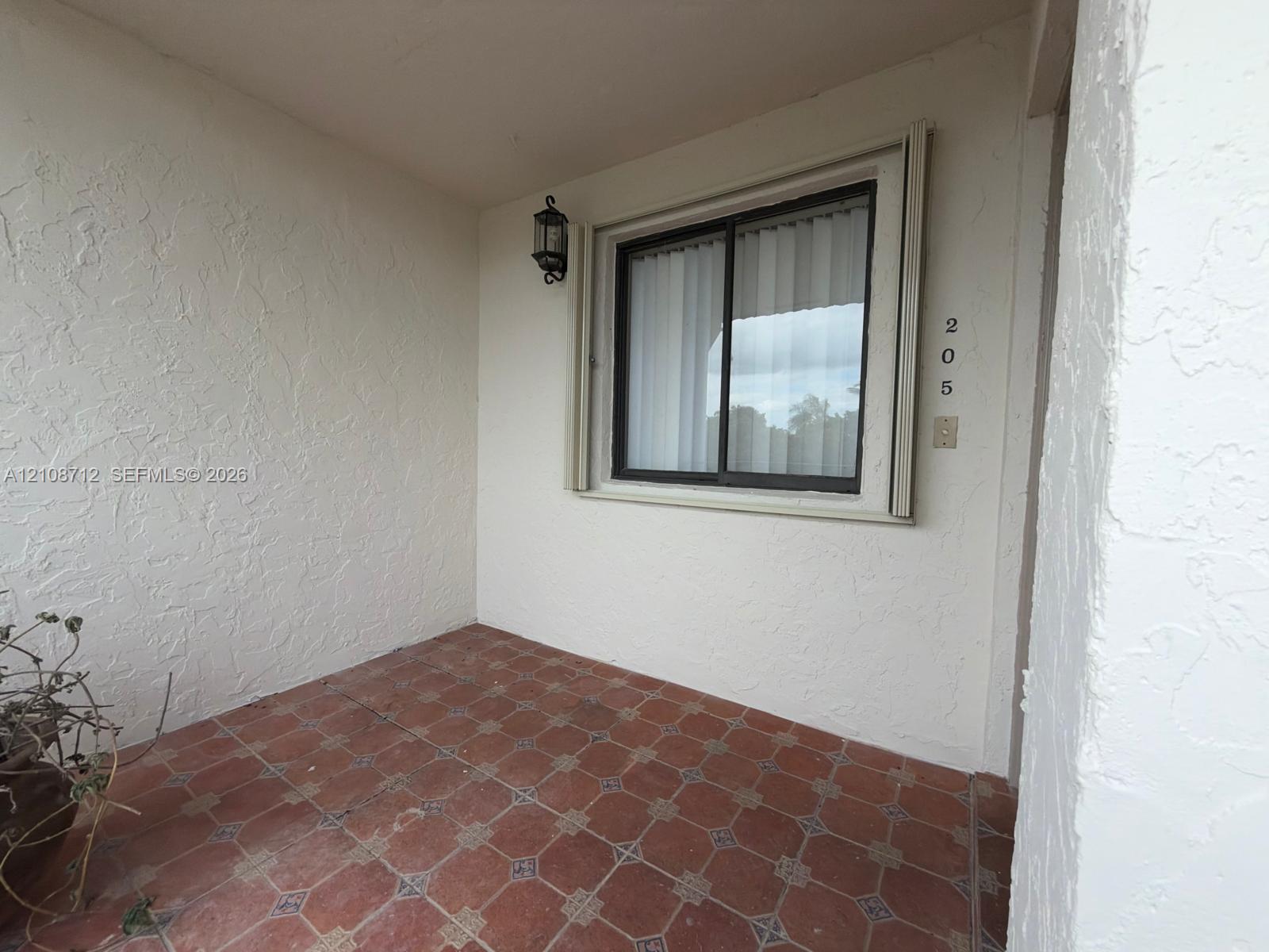 Covered entryway featuring terra cotta tile flooring, stucco wall finish, wall-mounted lantern lighting, and a window with a metal frame