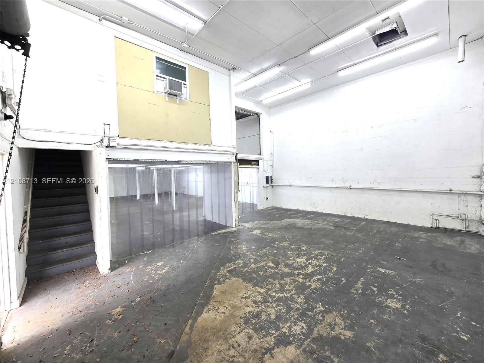 Industrial workspace featuring high ceilings, concrete flooring, overhead fluorescent lighting, and fixed staircase access to an elevated mezzanine level