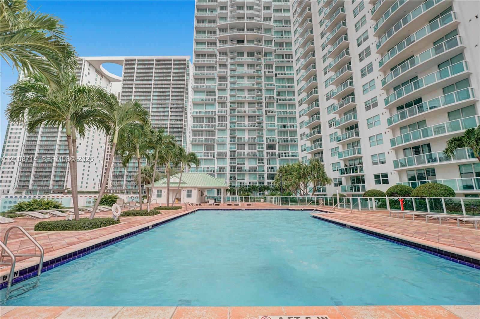 Swimming pool featuring tile edging, paved decking, and integrated metal handrails, set amidst palm trees and high-rise structures