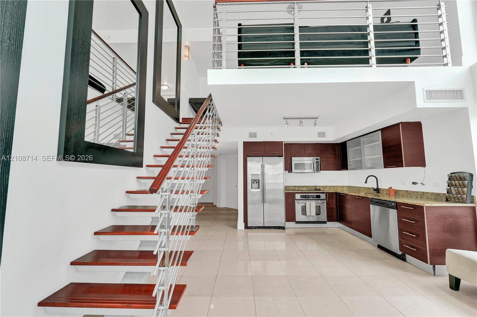 Open-concept interior featuring a stainless steel appliance suite, rich-toned cabinetry with stone-finish countertops, ceramic tile flooring, a mezzanine level with metal railings, and a floating staircase with wood-finish treads and metal cabling Open-concept interior featuring a stainless steel appliance suite, rich-toned cabinetry with stone-finish countertops, ceramic tile flooring, a mezzanine level with metal railings, and a floating staircase with wood-finish treads and metal cabling
