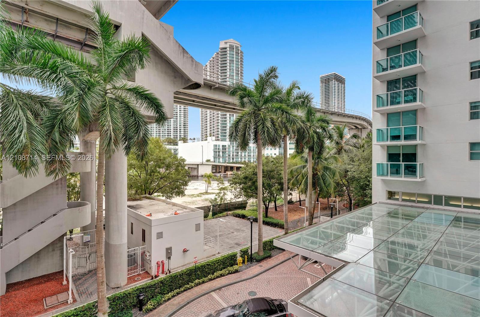 High-rise building with glass-paneled balconies and a glass-covered entryway featuring patterned paver driveway and mature palm landscaping