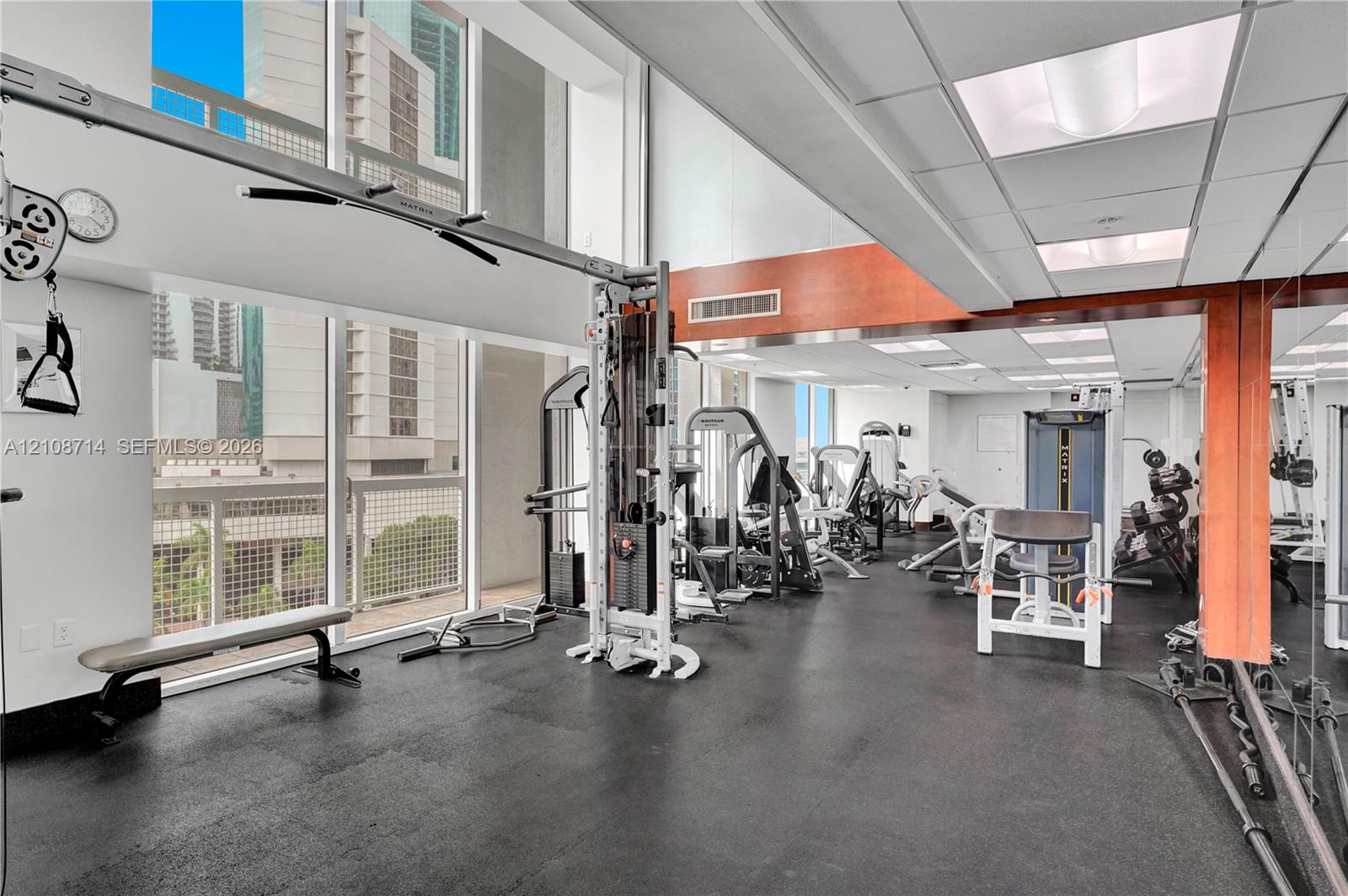 Fitness center featuring floor-to-ceiling windows, rubberized flooring, wood-paneled accents, mirrored walls, and drop-ceiling lighting