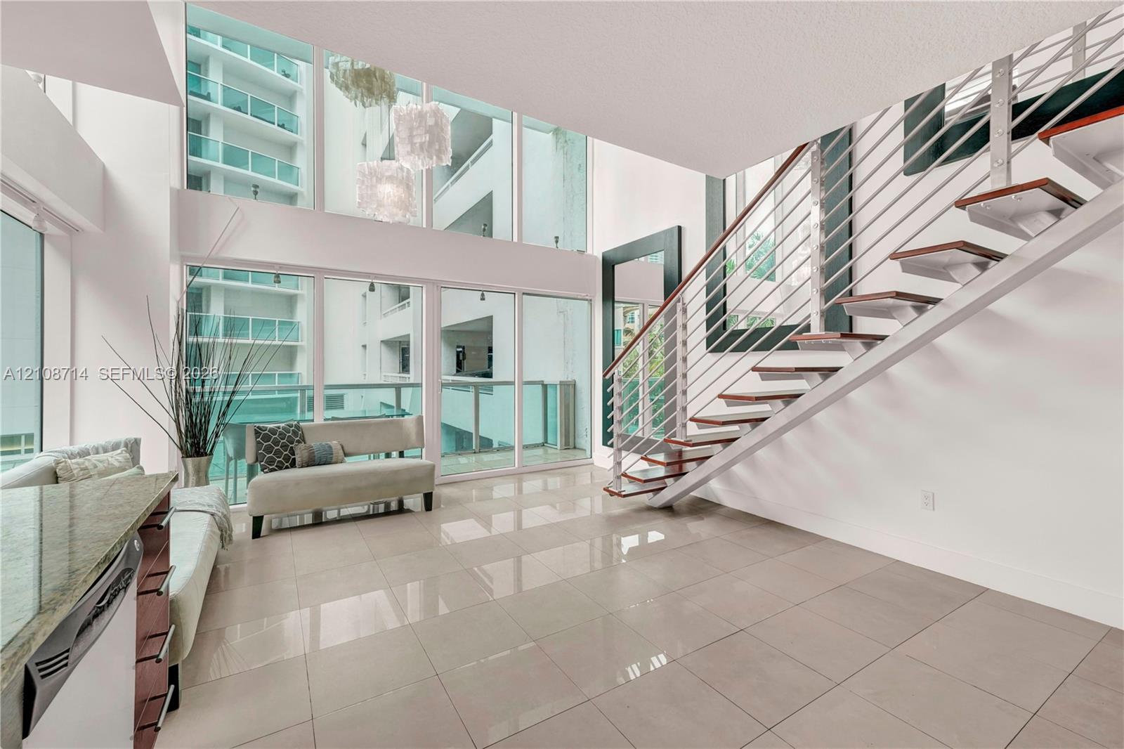 Double-height floor-to-ceiling windows, open-riser staircase with horizontal cable railing, neutral large-format tile flooring, and built-in stone-surface kitchen cabinetry Double-height floor-to-ceiling windows, open-riser staircase with horizontal cable railing, neutral large-format tile flooring, and built-in stone-surface kitchen cabinetry