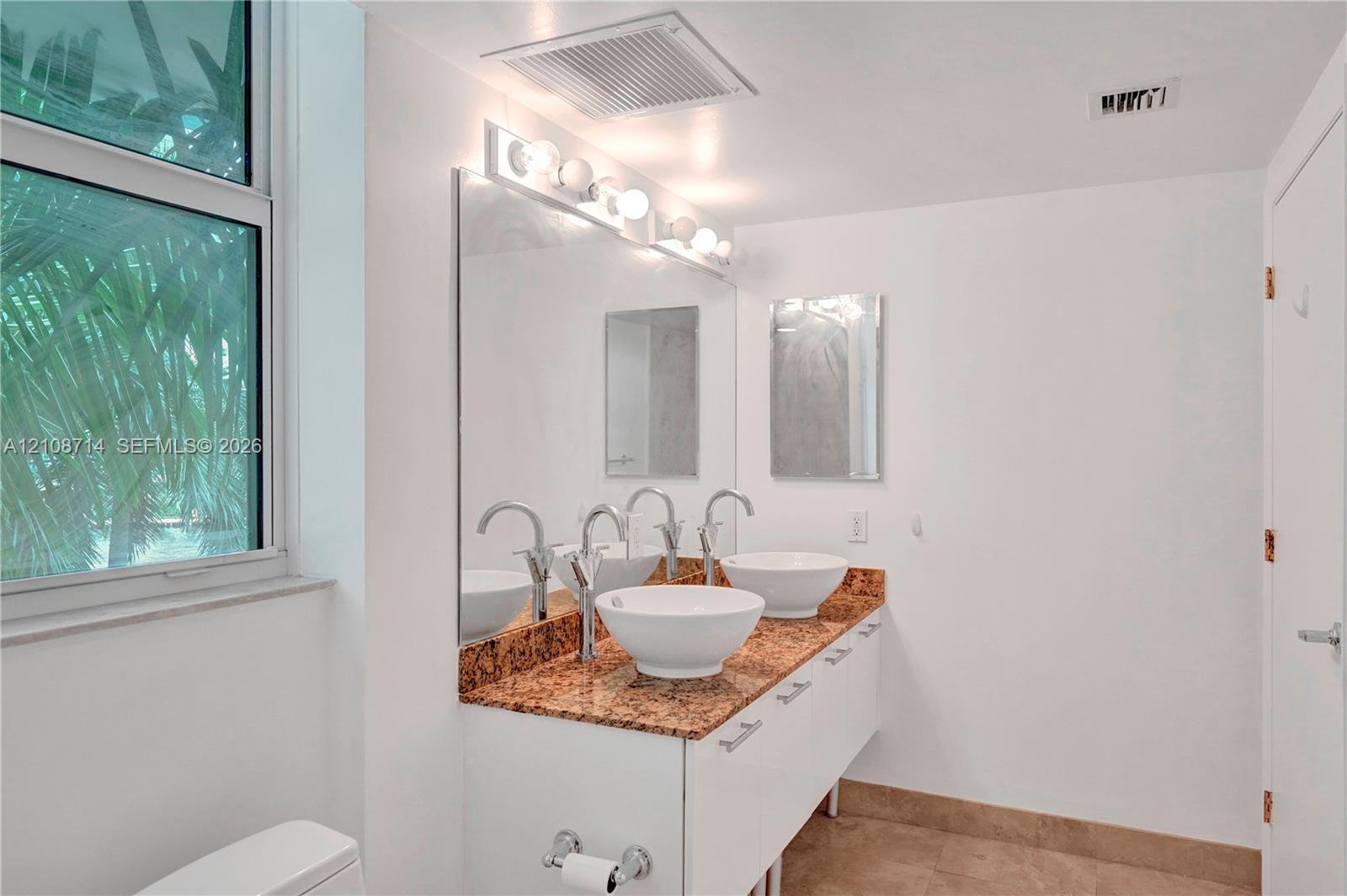 Double vanity featuring white cabinetry, stone-surface countertop, dual vessel sinks, and wall-mounted mirror with integrated lighting
