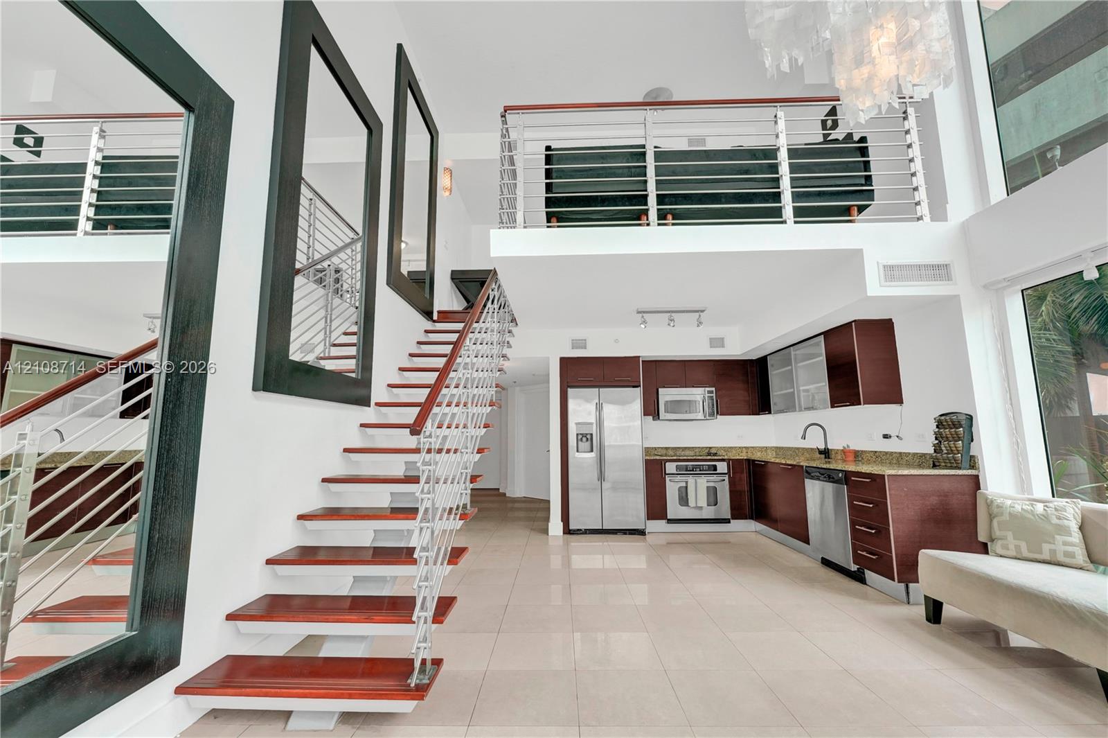 Open-concept interior featuring high ceilings, wood-tread staircase with cable railing, lofted level, tile flooring, and kitchen cabinetry with stainless steel appliances Open-concept interior featuring high ceilings, wood-tread staircase with cable railing, lofted level, tile flooring, and kitchen cabinetry with stainless steel appliances