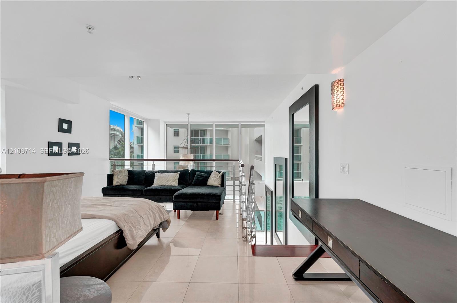 Open-concept loft area featuring large-format tile flooring, a built-in dark wood desk, floor-to-ceiling windows, and a metal-railing staircase