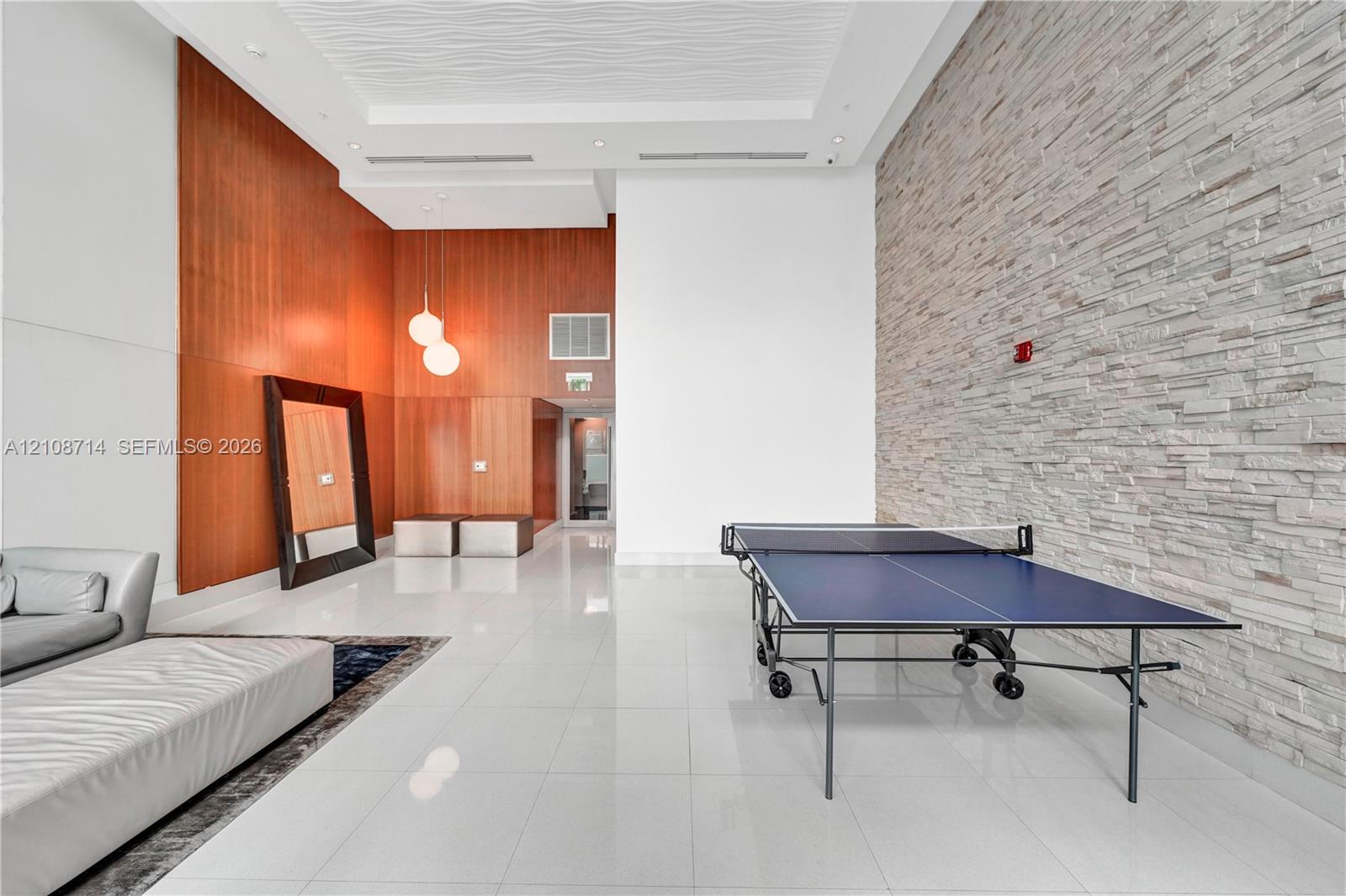 High-ceiling interior featuring smooth wood paneling, floor-to-ceiling textured stone accent wall, glossy large-format tile flooring, recessed lighting, and modern globe pendant fixtures