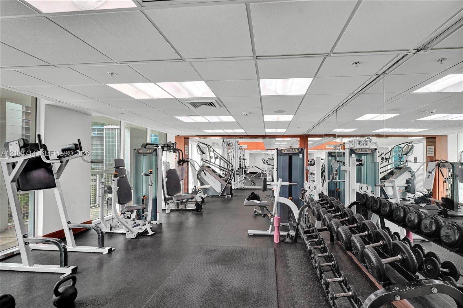 Fitness facility featuring rubberized flooring, expansive wall-to-wall mirrors, and a suspended drop ceiling with recessed lighting