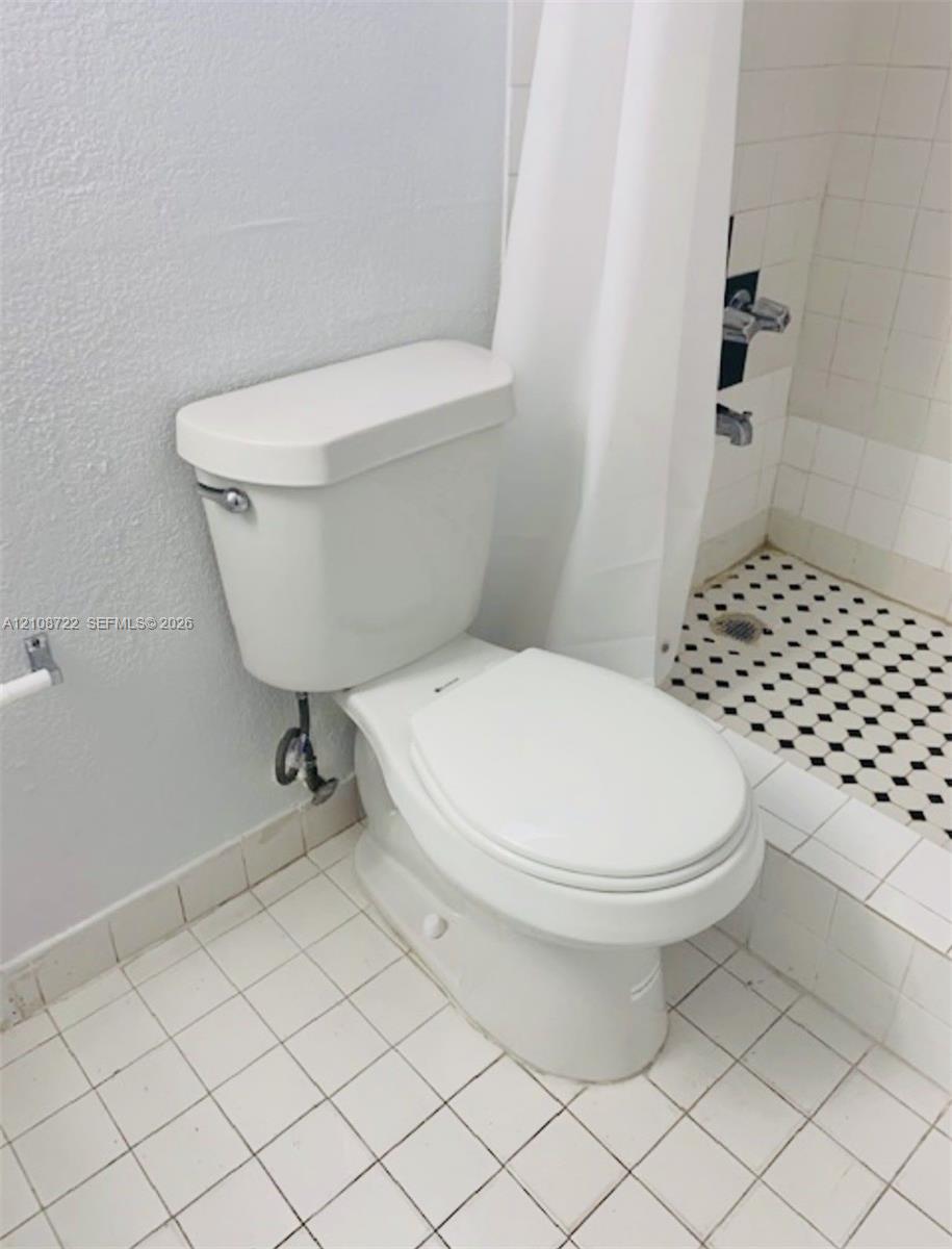 Bathroom featuring square white floor tile, a porcelain toilet, and a walk-in shower with black-and-white patterned mosaic flooring and a metal wall-mounted faucet assembly Bathroom featuring square white floor tile, a porcelain toilet, and a walk-in shower with black-and-white patterned mosaic flooring and a metal wall-mounted faucet assembly