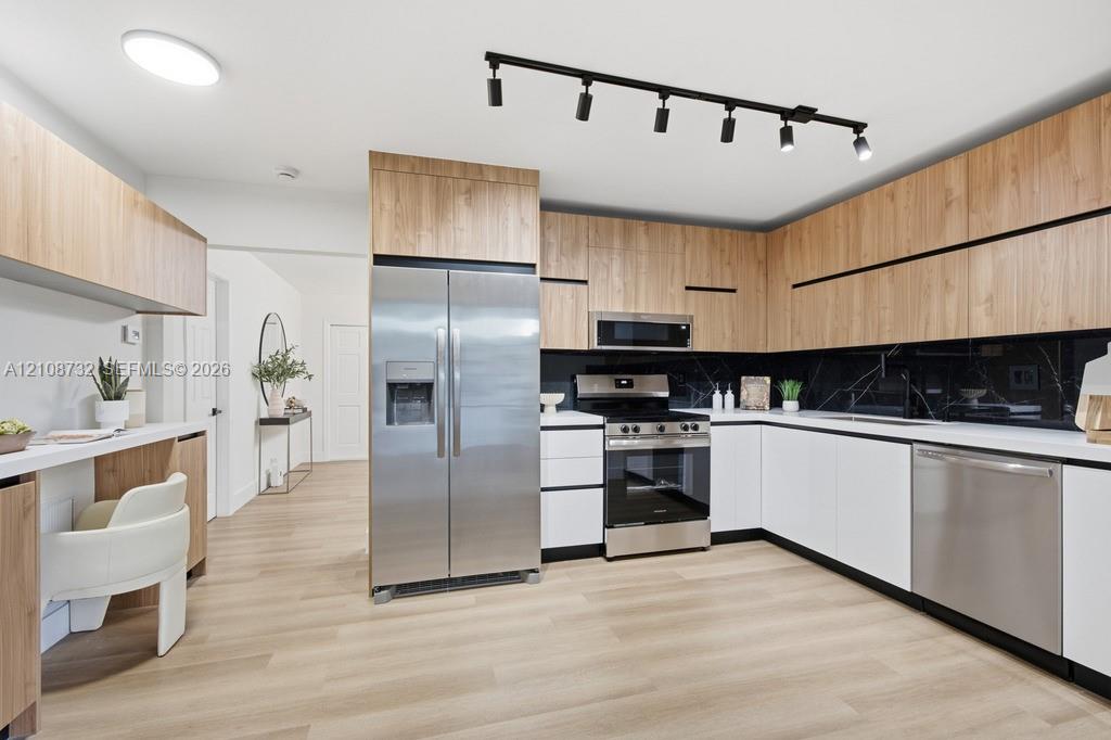 Kitchen featuring flat-panel wood-finish cabinetry, stainless steel appliances, dark stone-finish backsplash, built-in desk area, and matte black track lighting