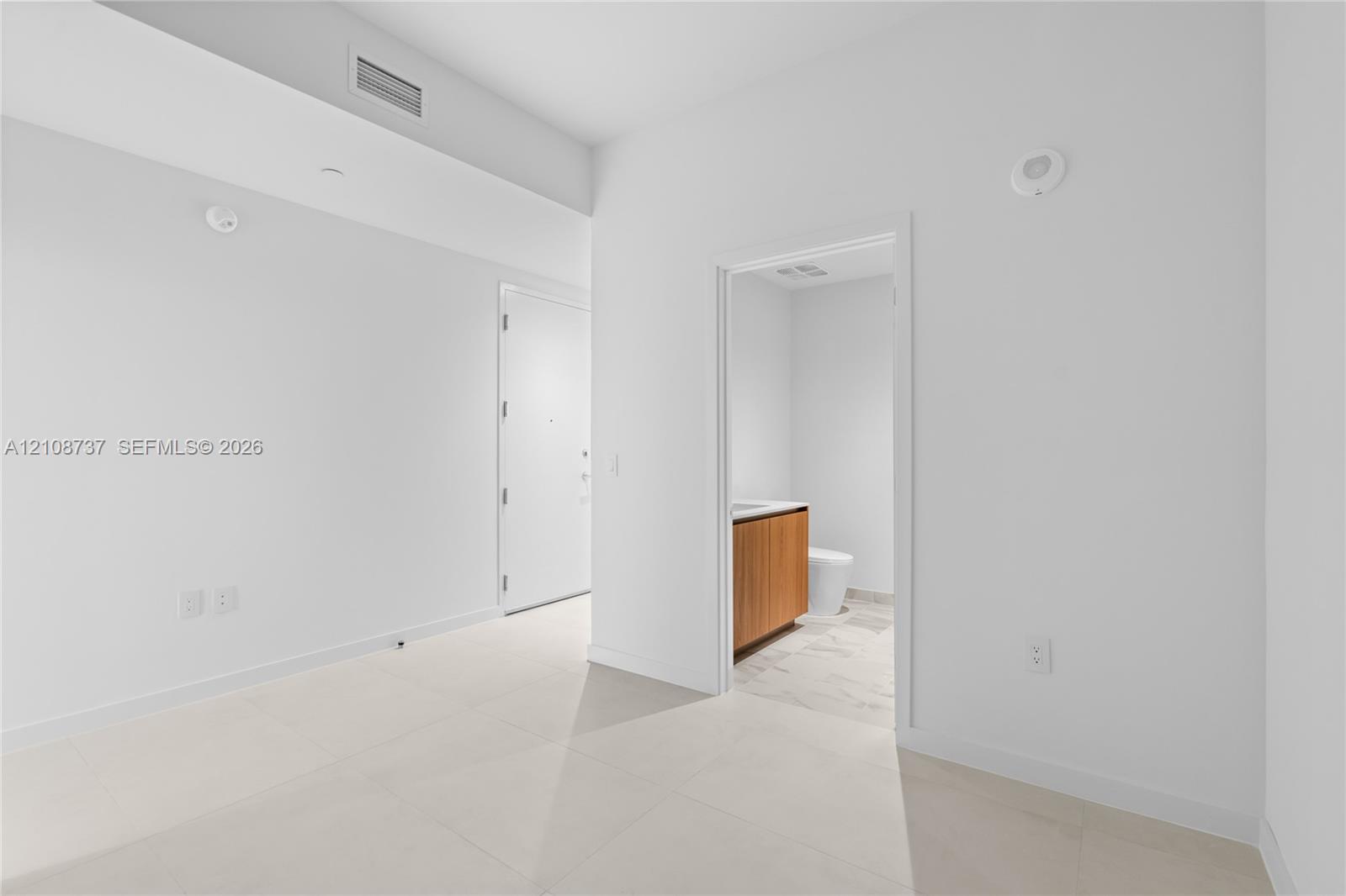 Interior space featuring large-format tile flooring, neutral wall finishes, and a door leading to a bathroom equipped with a wood-finish vanity cabinet