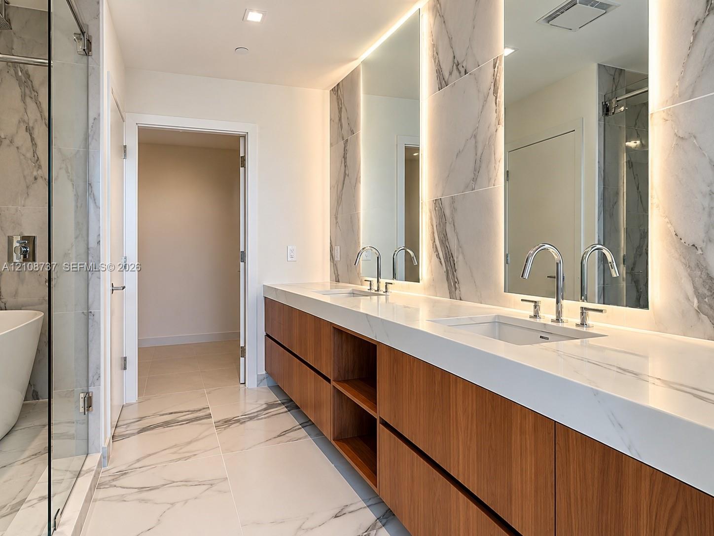 Floating wood-finish vanity with dual sinks, white stone countertop, back-lit mirrors, and floor-to-ceiling stone tile throughout