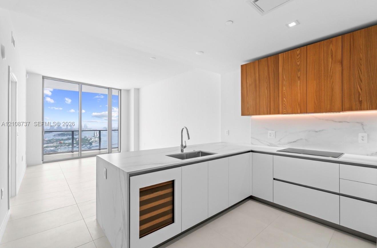Modern kitchen featuring stone-surfaced countertops, custom wood-finish cabinetry, built-in wine refrigeration, integrated cooktop, and floor-to-ceiling windows