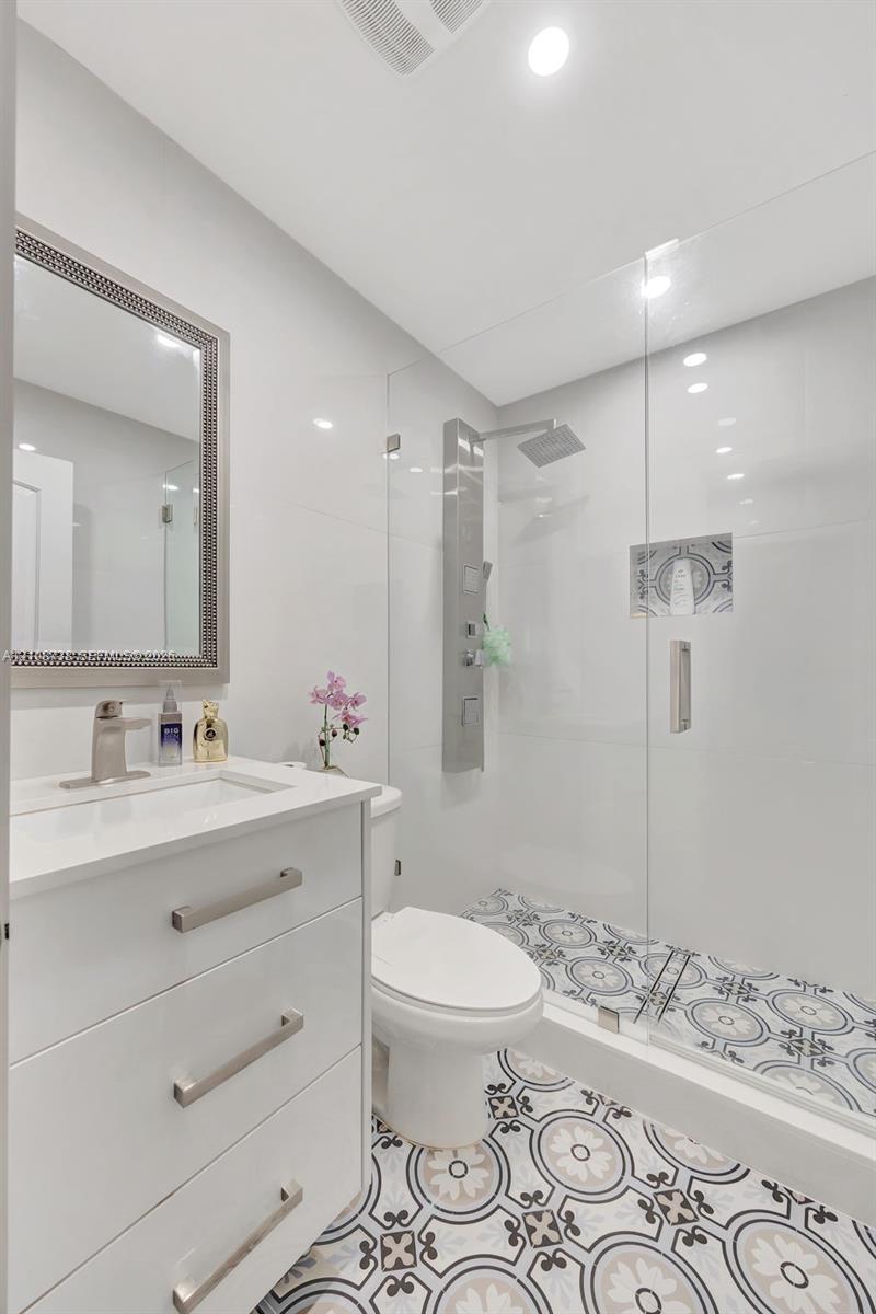 White vanity with a rectangular sink, brushed nickel hardware, patterned tile flooring, walk-in shower with a glass enclosure, and a wall-mounted shower panel