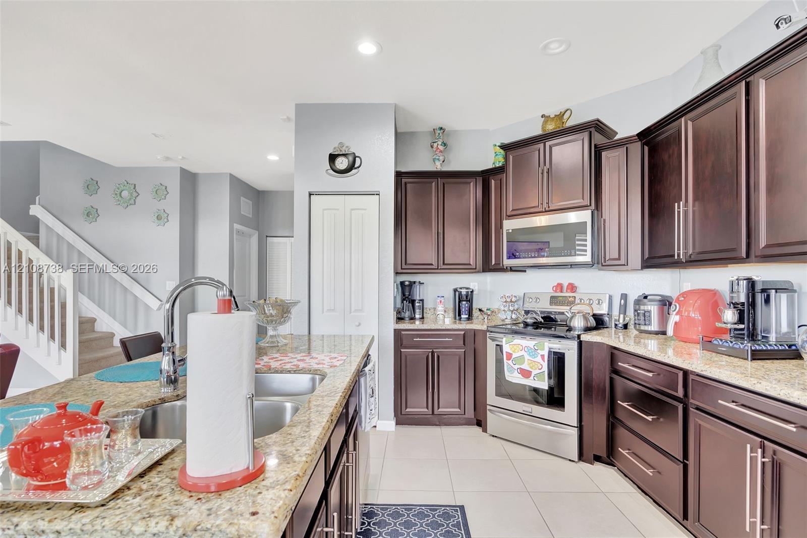 Kitchen featuring dark wood cabinetry, stone-surface countertops, stainless steel appliances, a center island with an integrated double-basin sink, and tile flooring