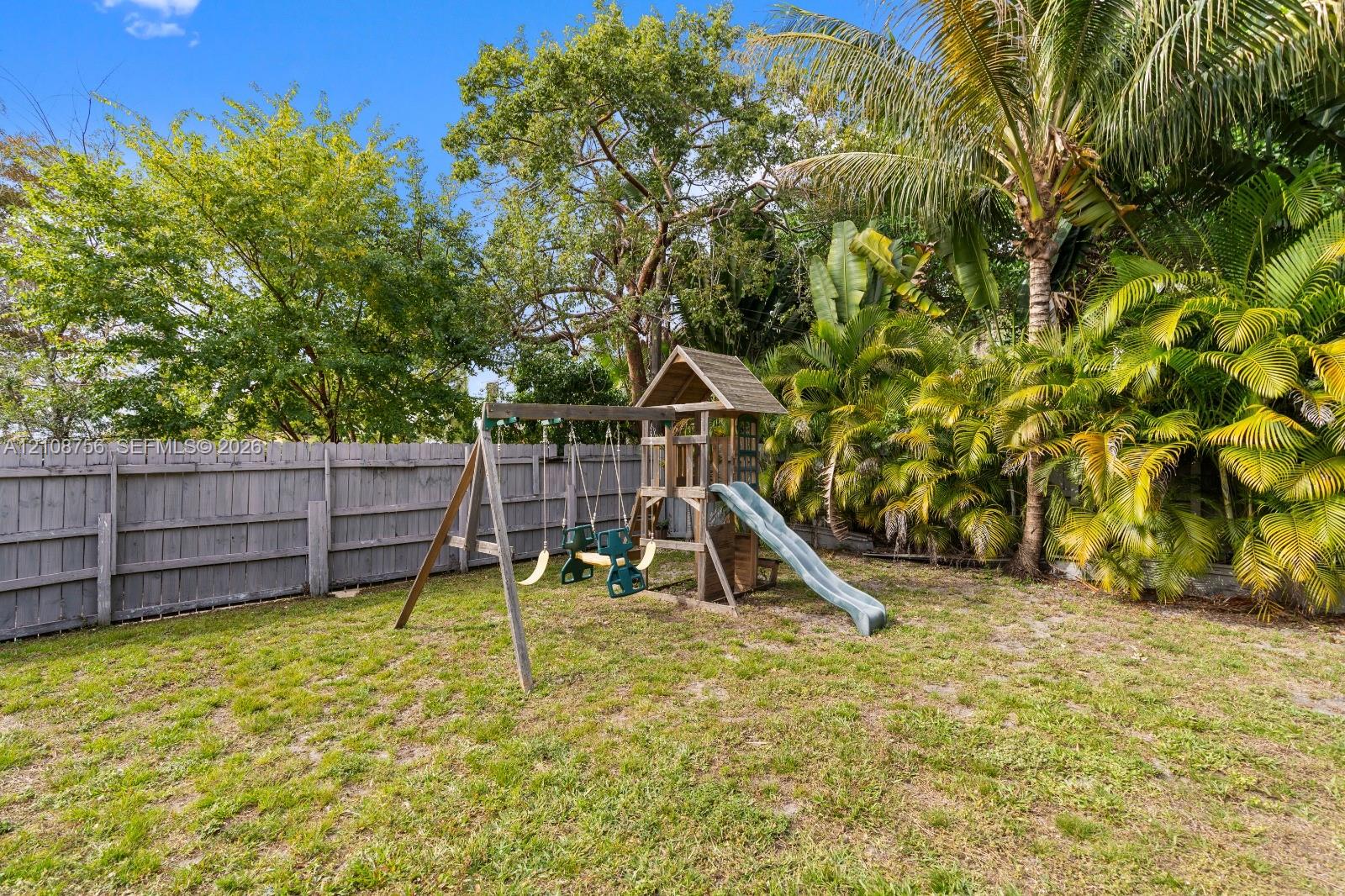 Fenced backyard featuring mature trees, tropical palms, and a wooden play structure