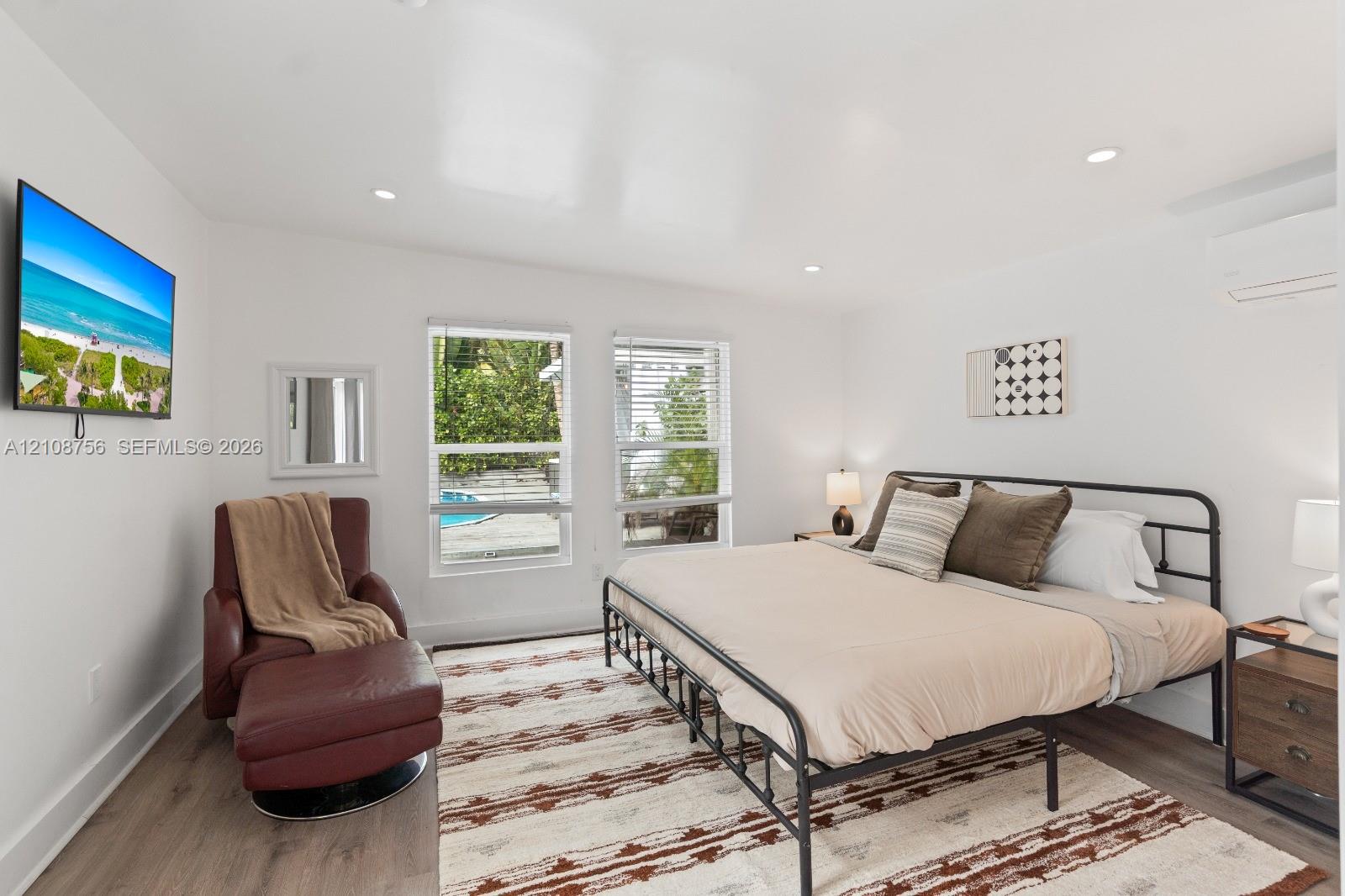 Neutral-toned room featuring wood-finish flooring, recessed lighting, dual windows with blinds, and a wall-mounted climate control unit