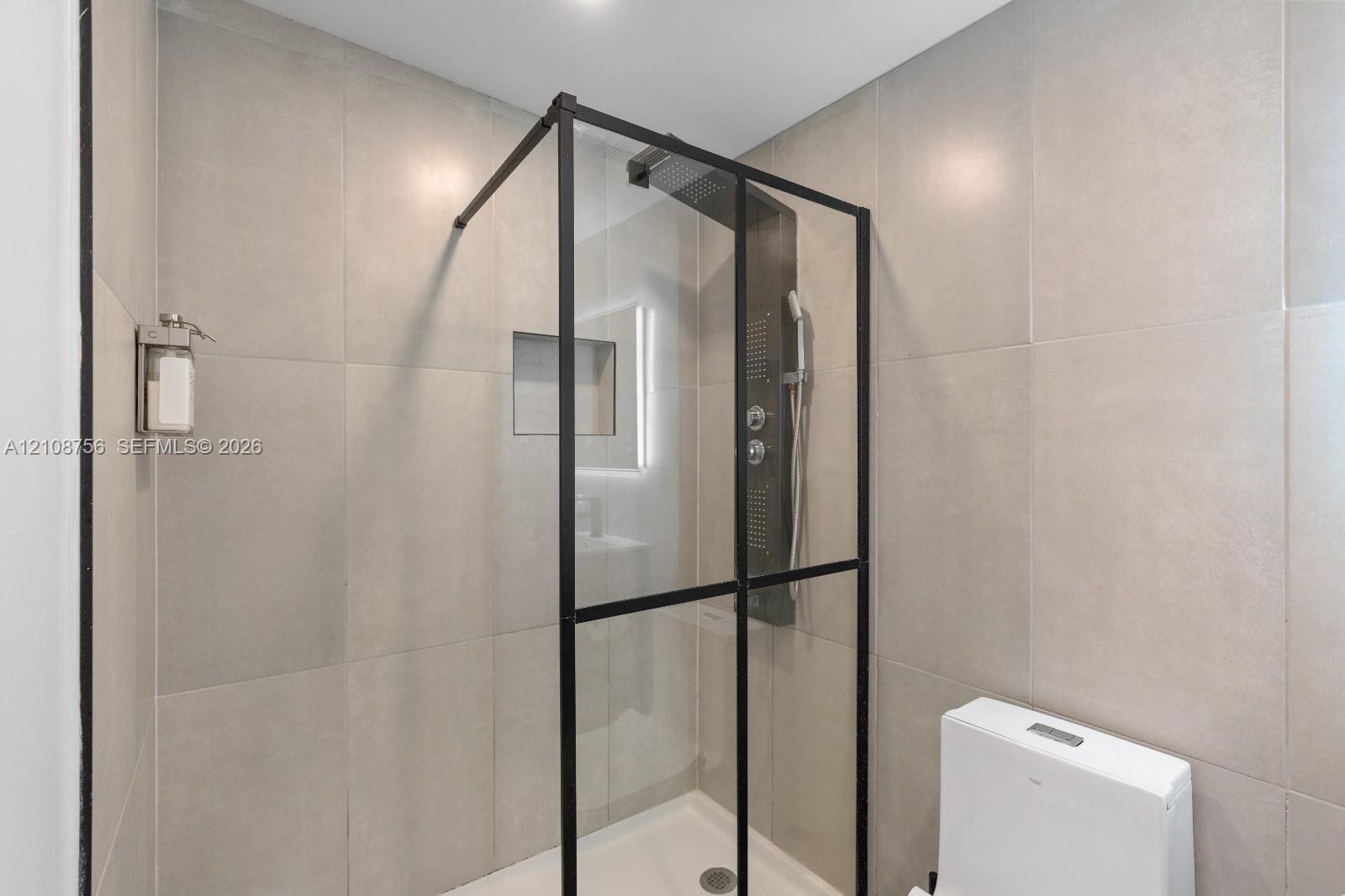Walk-in shower enclosure featuring matte black framing, floor-to-ceiling large-format tile surround, integrated shower panel with handheld sprayer, recessed wall niche, and low-profile white base