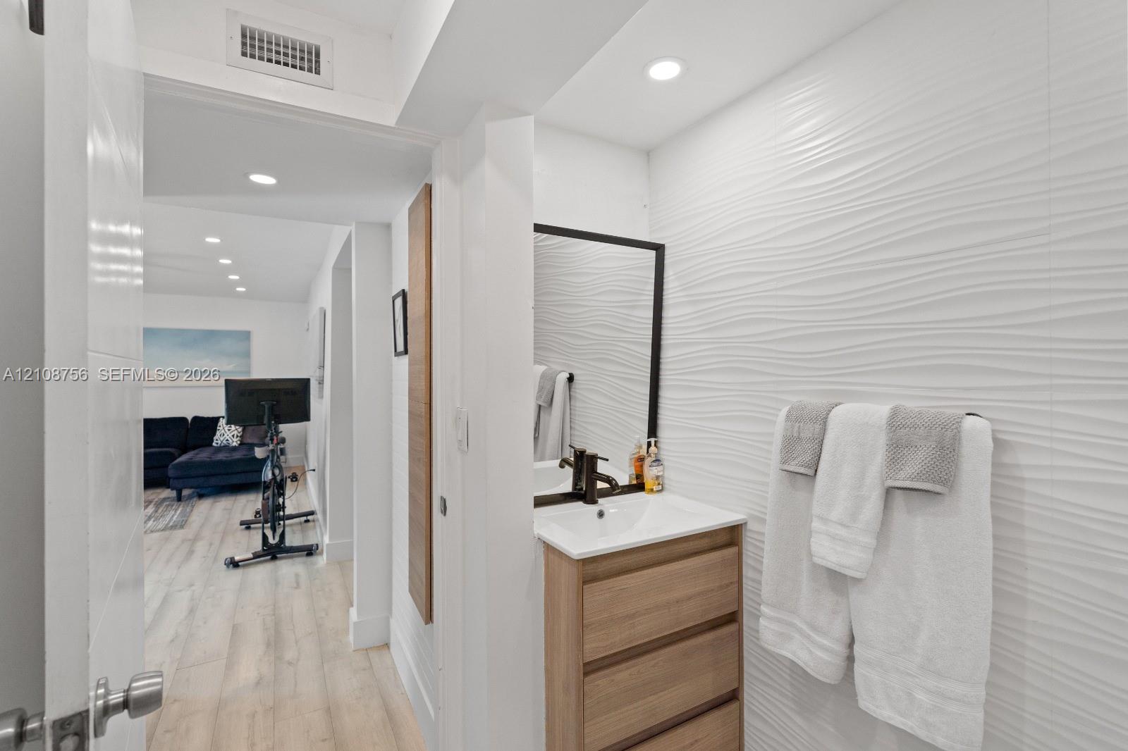 Modern vanity with wood-finish cabinetry, white countertop, textured wave-pattern accent wall, and framed rectangular mirror
