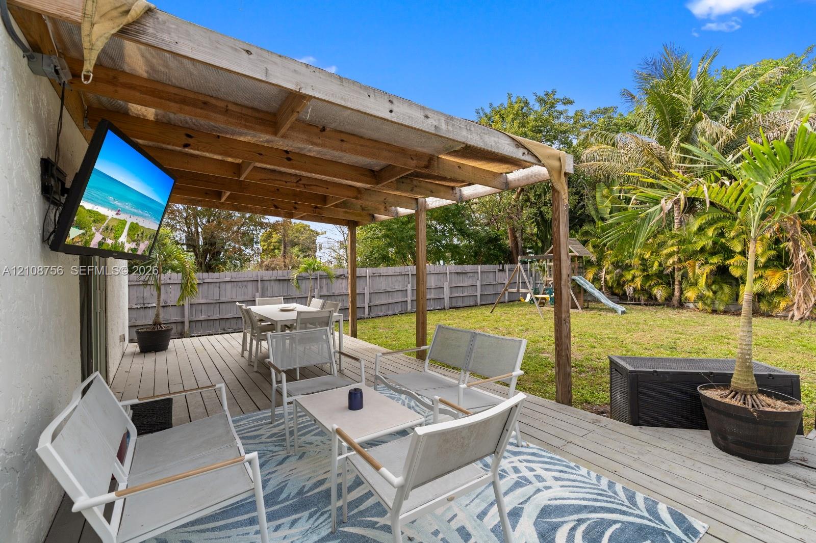 Covered wooden deck featuring a mounted television and wood-beam construction, complemented by a fenced backyard