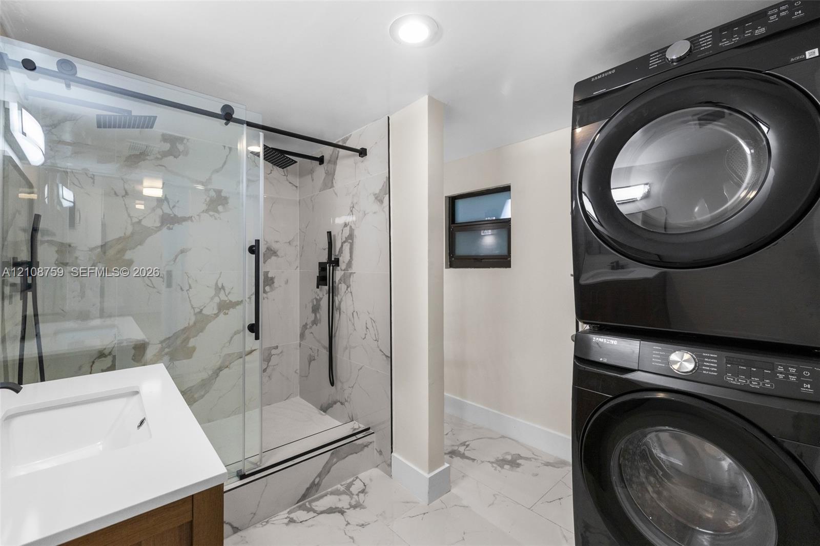 Walk-in shower with sliding glass door, stone-finish surround, and matte black fixtures, paired with a modern vanity featuring a rectangular integrated sink and a stacked washer-dryer unit