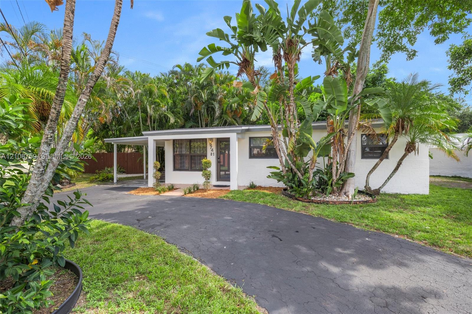 White painted brick exterior featuring a covered carport, asphalt driveway, and mature tropical landscaping White painted brick exterior featuring a covered carport, asphalt driveway, and mature tropical landscaping