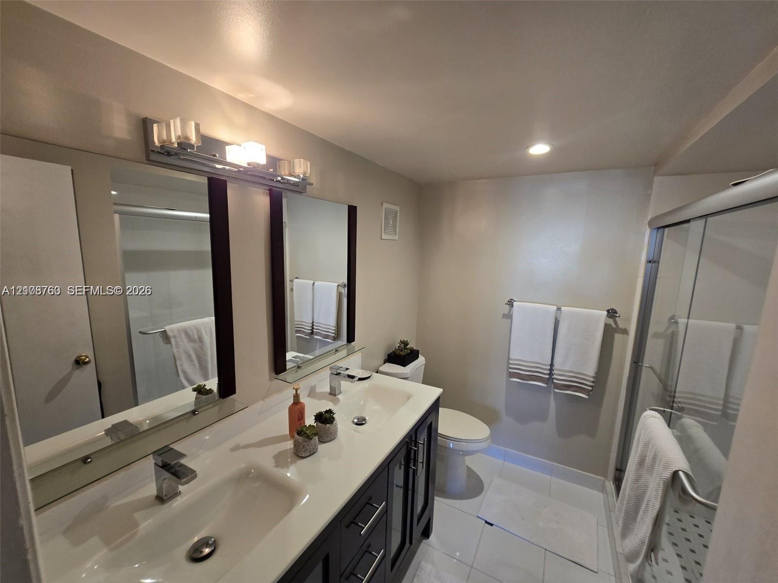 Double-sink vanity featuring dark wood-finish cabinetry, a solid-surface countertop, and chrome fixtures, paired with a glass-enclosed shower, dual-frame mirrors, and polished tile flooring