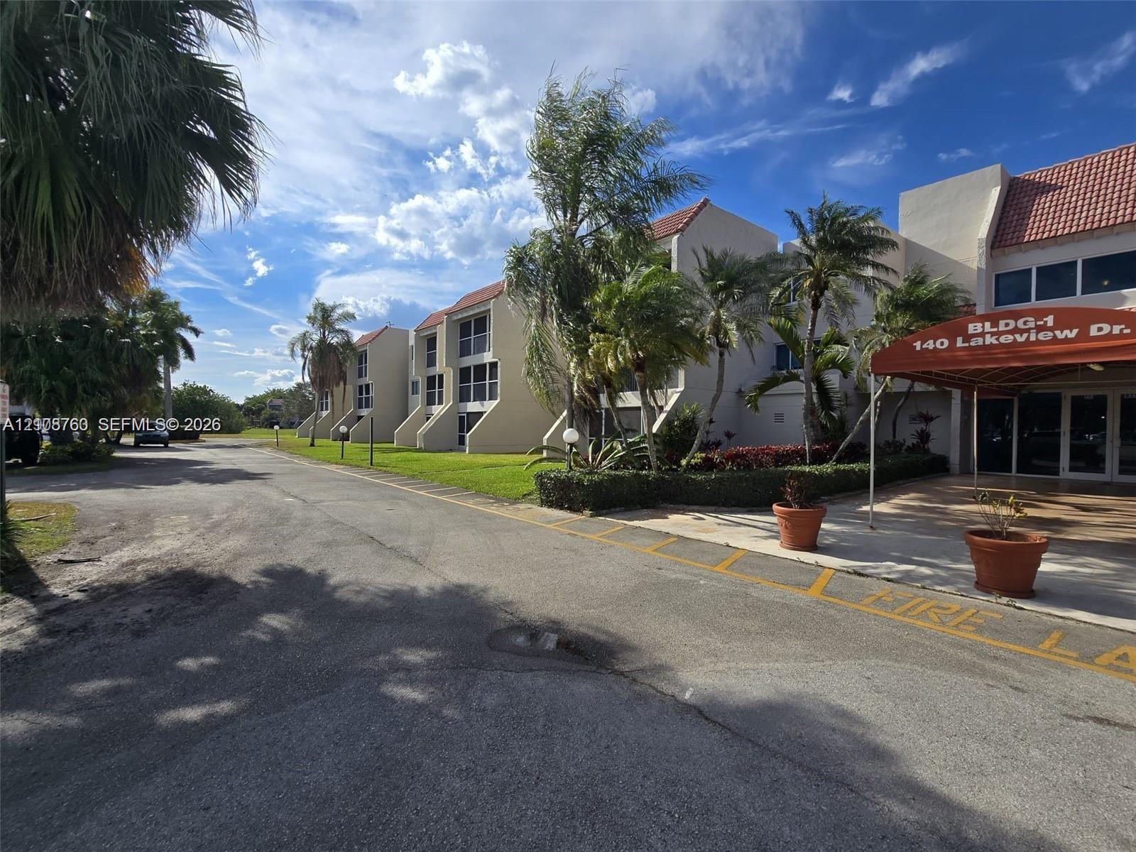 Multi-story exterior featuring a light-toned stucco faade, terra-cotta-hued roof tiles, and ground-level covered entryway with a red fabric awning, complemented by landscaped perimeter gardens and mature palms