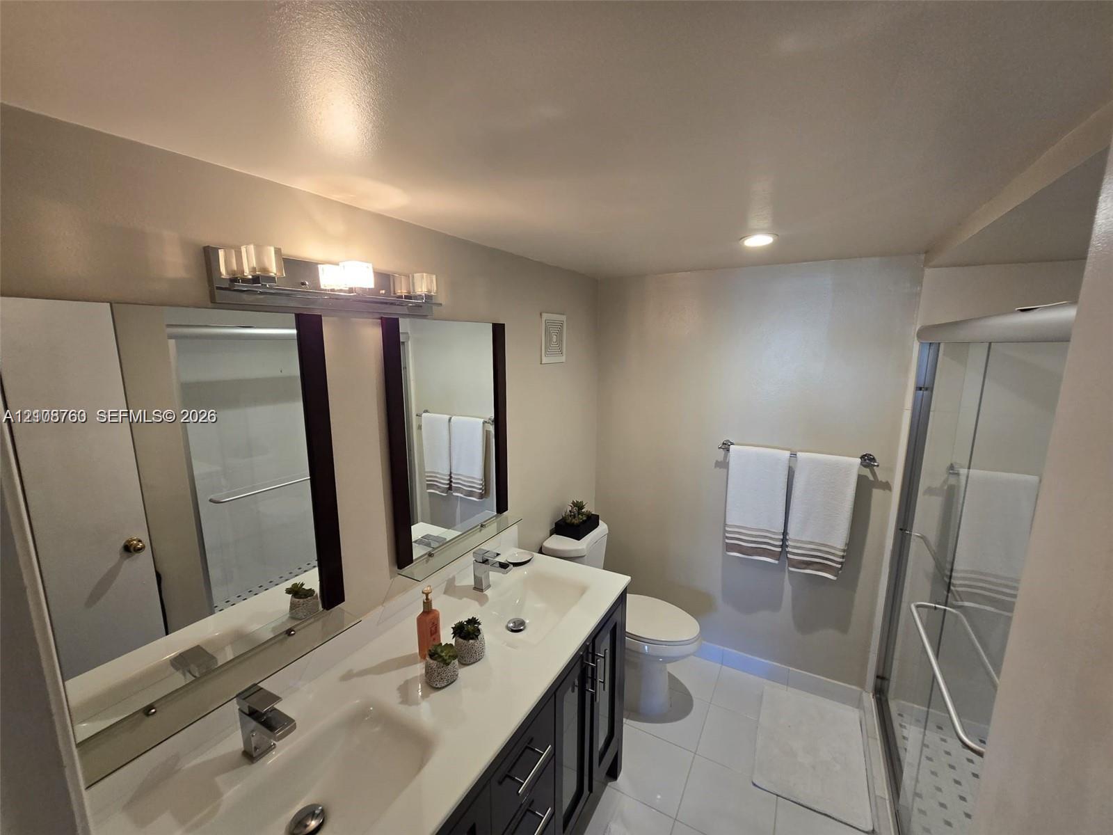 Dual-sink vanity with a solid-surface countertop, dark cabinetry, full-width mirror, and a walk-in glass-enclosed shower featuring tiled flooring