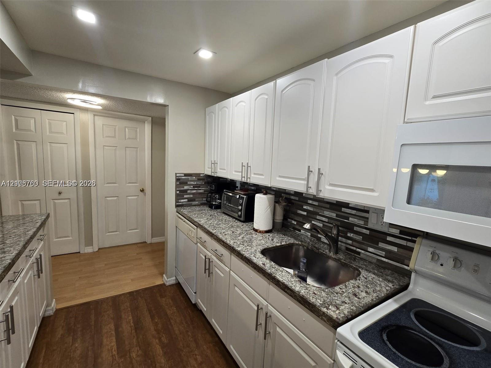 Kitchen featuring white shaker-style cabinetry, multi-tonal linear tile backsplash, stone-finish countertops, stainless steel undermount sink with gooseneck faucet, and wood-finish flooring