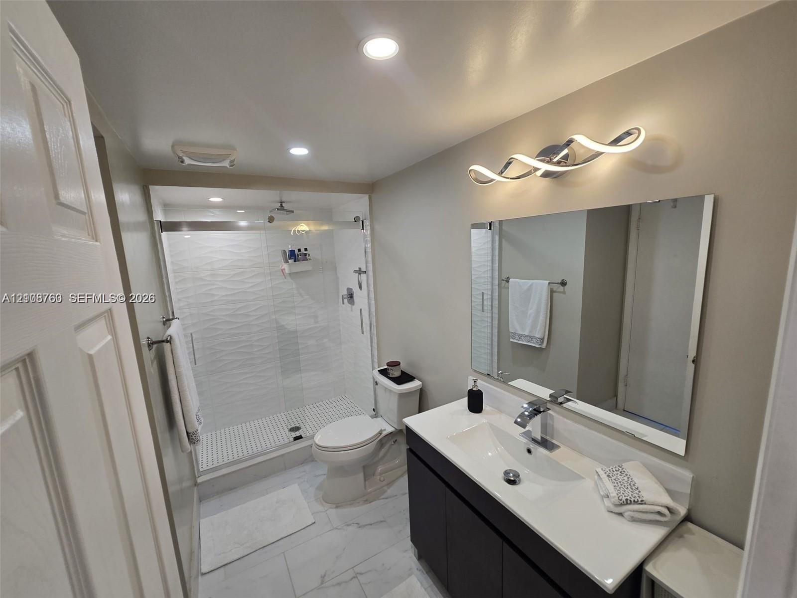 Bathroom featuring a dark wood-finish vanity with an integrated countertop sink, walk-in shower with textured wall tile, frameless glass enclosure, and contemporary wave-style wall lighting