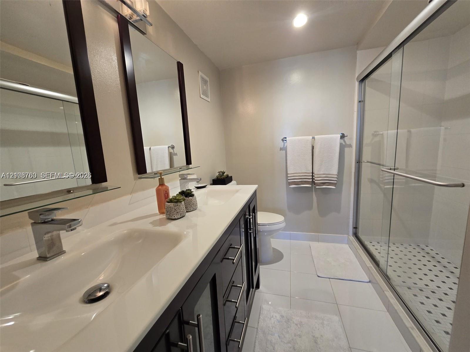Dual-sink vanity with a solid-surface countertop, dark wood cabinetry, rectangular glass-enclosed shower, and polished chrome fixtures